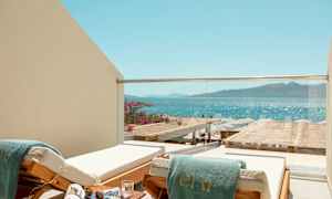 En solstol med en grön handduk vid en balkong med glasräcke, med utsikt över havet och en strand med parasoller. (AI-genererad text)