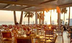 Uteservering med bord och stolar vid solnedgången, med palmer och hav i bakgrunden. (AI-genererad text)