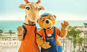 Två maskotar, en giraff och en björn, står framför ett hav i bakgrunden. (AI-genererad text)