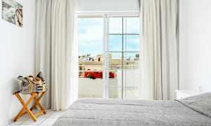 Ett ljust sovrum med en grå bäddad säng, en trästol med en ihoprullad väska, gardiner, och en balkongdörr som leder till en utsikt med blommor och byggnader. (AI-genererad text)