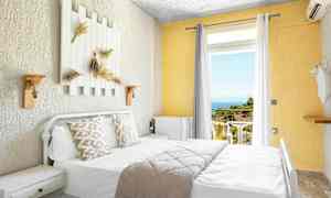 Ljust hotellrum med säng, dekorerade kuddar och utsikt mot balkong med hav och grönska. (AI-genererad text)