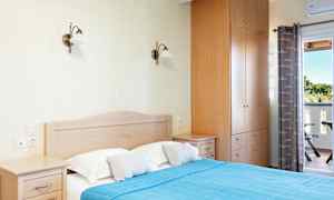 Ett sovrum med en dubbelsäng som har en blå överkast och två kuddar, en garderob och nattduksbord. En utomhus balkong och gardiner syns i bakgrunden. (AI-genererad text)