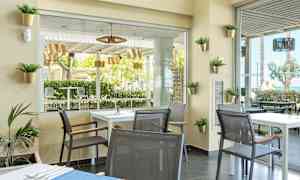 Ett modernt inrett café med bord och stolar, utsikt mot grönskande träd och utemöbler. (AI-genererad text)