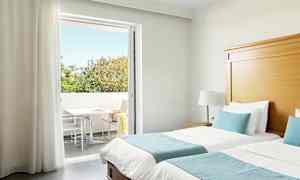 Hotellrum med två enkelsängar, blå sängkläder och en direkt utgång till en balkong med träd som syns i bakgrunden. (AI-genererad text)