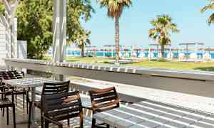 En terrass med bord och stolar, med utsikt över en grön gräsmatta, palmer och stranden i bakgrunden. (AI-genererad text)