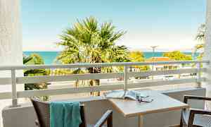 En balkong med utsikt över en palm, havet och grannbyggnader. (AI-genererad text)