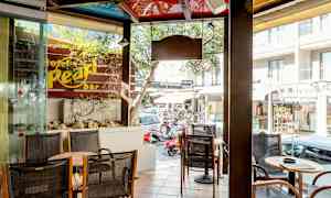 Ett caféområde med sittplatser och bord bredvid ett skyltfönster. Utomhus syns en gata med butiker och bilar. (AI-genererad text)