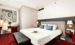 Ett hotellrum med en stor säng, två blå kuddar, ett skrivbord med stol, röd matta och moderna lampor. (AI-genererad text)