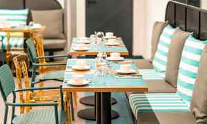 Bild av en restaurang med bord dukade med koppar, glas och bestick. Stolar och bänksittplatser med randiga kuddar syns. (AI-genererad text)