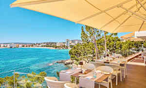 Uteservering med utsikt över en klarblå havsvik och byggnader i fjärran. (AI-genererad text)