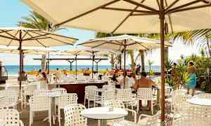 Utomhusrestaurang med vita bord och stolar under parasoller, utsikt mot havet. (AI-genererad text)