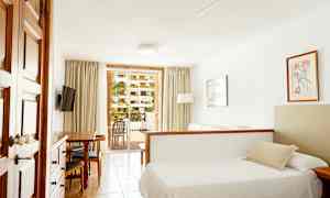 Ett hotellrum med en säng, soffa och matbord, samt utsikt mot en balkong med grönska utanför. (AI-genererad text)