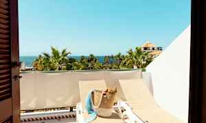 Utsikt över en solig balkong med solstolar, en korg väska, palmer och havet i bakgrunden. (AI-genererad text)