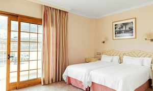 Ett hotellrum med två enkelsängar med vita sängkläder, en tavla på väggen och en glasdörr med gardiner. (AI-genererad text)