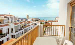 Utsikt från en balkong med ett bord och stolar över vita byggnader med röda tak och havet i bakgrunden. (AI-genererad text)