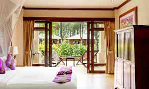 Ett sovrum med en säng och kuddar i lila färger, utsikt mot en trädgård genom stora fönster med dörrar. (AI-genererad text)