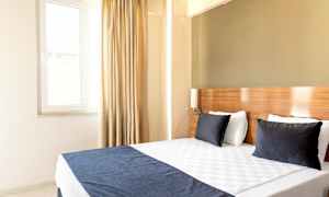 Ett ljust hotellrum med en dubbelsäng, två mörka kuddar och ett fönster med gardiner. (AI-genererad text)