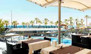 Utsikt från en uteservering mot en poolområde med parasoller och palmer samt havet i bakgrunden. (AI-genererad text)