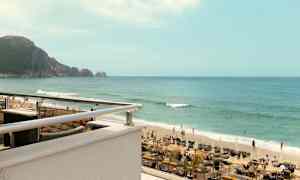 En bild av en strand med solsängar, parasoller och människor som simmar i havet. I bakgrunden syns en bergsklippa. (AI-genererad text)