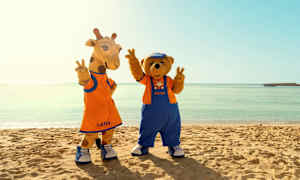 Två maskotkaraktärer med namnen Lollo och Bernie står på en strand framför klara havet. (AI-genererad text)