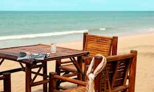 Stolar och bord av trä står på en strand med havet i bakgrunden. (AI-genererad text)
