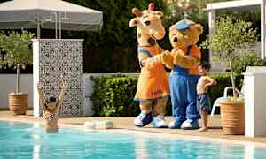 Två maskotkaraktärer, en giraff och en björn, står bredvid poolen utomhus. (AI-genererad text)