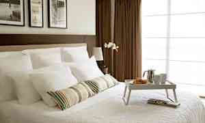 Ett rum med en bäddad dubbelsäng, vit sängklädsel, och en frukostbricka med croissanter, en kaffekanna och koppar. I bakgrunden syns bruna gardiner och tavlor på väggen. (AI-genererad text)