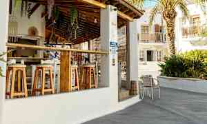 En utomhusbar med trästolar och tak av terrakottaplattor. En palm och byggnader syns i bakgrunden. (AI-genererad text)