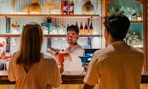 Två personer sitter på en bänk vid en bardisk, som är dekorerad med flaskor och färgstarka detaljer. (AI-genererad text)