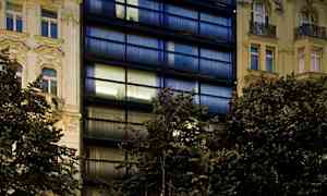 Bilden visar en modern byggnad med mörka glasfasader och klassiska byggnader på vardera sida, med en restaurang och parkering i förgrunden. (AI-genererad text)