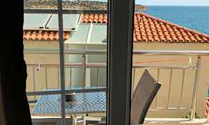 Utsikt från ett rum mot en balkong med bord och stolar, tak med röda takpannor och havet i bakgrunden. (AI-genererad text)