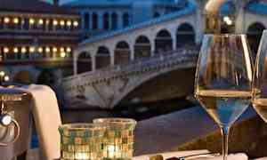 En tallrik med skaldjur och ett glas vitvin på ett bord med utsikt över en upplyst bro i skymningen. (AI-genererad text)