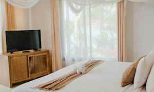 Ett ljust sovrum med en dubbelsäng med vitt sängkläder, en TV på en träbyrå, stora fönster och gardiner samt dekorativa taktyger. (AI-genererad text)