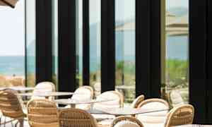 En uteservering med bord och stolar, intill en byggnad med stora glasfönster. (AI-genererad text)