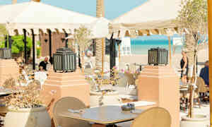 Uteservering med bord och stolar under parasoll nära stranden. (AI-genererad text)