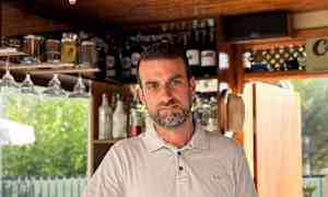 En vit skjorta vid en bar med hyllor fyllda av flaskor i bakgrunden, samt en cocktailglas med en färgad dryck framför sig. (AI-genererad text)