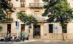 Bild av en byggnad med fasad av ljus sten. Byggnaden har flera våningar med balkonger, omgivna av gröna träd. (AI-genererad text)