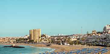 Sandstrand med många blå parasoller, havet och byggnader i bakgrunden. (AI-genererad text)