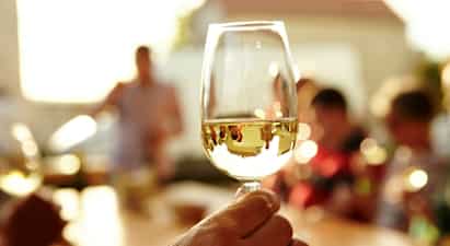 En person håller i ett glas vitt vin med en solbelyst bakgrund med människor. (AI-genererad text)