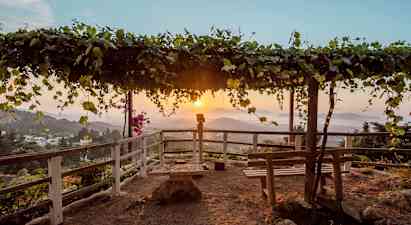 En utkiksplats med en träpergola täckt av löv samt bänkar och staket runtom. Solnedgång över ett bergigt landskap i bakgrunden. (AI-genererad text)