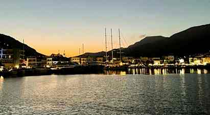 En hamn i skymningen med båtar förtöjda vid en kaj och upplysta byggnader på avstånd. Berg syns i bakgrunden. (AI-genererad text)