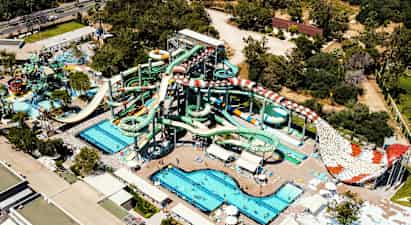 Ett vattenland med flera stora vattenrutschbanor, pooler och solstolar, omgiven av gröna träd och byggnader. (AI-genererad text)