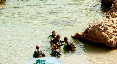 Grupp av personer i våtdräkter och dykarutrustning står i klart vatten intill klippor. (AI-genererad text)