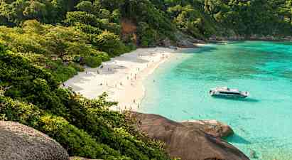 En sandstrand med människor, omgiven av grönskande träd och klart turkost vatten. En båt ligger nära stranden. (AI-genererad text)