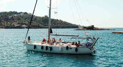 En segelbåt med flera personer ombord seglar nära en kustlinje med gröna kullar och några hus. (AI-genererad text)