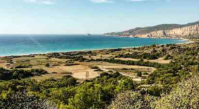En landskapsbild med gröna fält, buskar och berg som sträcker sig mot en strand med turkost hav under en klar himmel. (AI-genererad text)