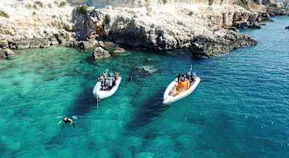 Två båtar ligger för ankar i klart turkost vatten nära en klippig kust. Dykare är i vattnet. (AI-genererad text)