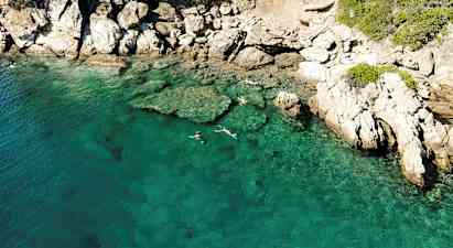 Klart turkost vatten vid en klippig kust, där personer snorklar nära ytan. (AI-genererad text)