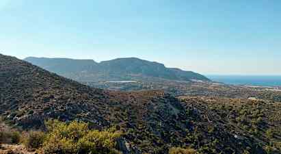 En vy över ett bergigt landskap med varierande växtlighet och havet i bakgrunden under en klarblå himmel. (AI-genererad text)