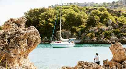 En segelbåt ligger förtöjd vid en rofylld vik omgiven av klippor och skog med en person som fiskar från stranden. (AI-genererad text)